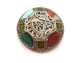 May include: A round silver-toned brooch featuring an intricate central knotwork design. The piece is accented with a light blue turquoise-colored stone, a dark orange carnelian-colored stone, a green malachite-colored stone, and two small square olive-green stones.