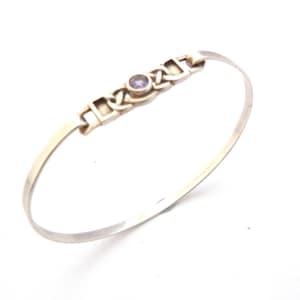 May include: A silver-toned bangle bracelet with a central decorative element. The design features a series of interlocking shapes and a small, round, purple gemstone. The bracelet has a simple, elegant design.