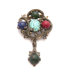 Op de afbeelding: Vintage broche met een sierlijk goudkleurig metalen filigraan ontwerp. De broche heeft een cluster van kleurrijke edelstenen, waaronder blauw, turquoise, rood en groen, met een hangende groene steen aan de onderkant. Een uniek sieraad.