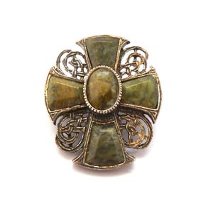 May include: A cross-shaped brooch featuring a central oval stone and four trapezoidal stones in shades of mottled olive green and brown. The metal frame has an antiqued silver finish with intricate openwork knot patterns between the arms.