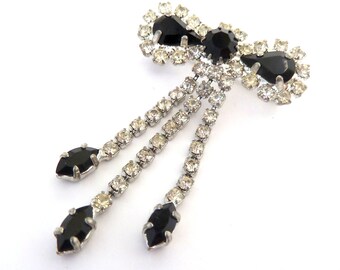 May include: A bow-shaped brooch featuring black teardrop and round stones surrounded by clear rhinestones. Three dangling chains extend from the center, each ending in a black marquise-shaped stone, all set in a silver-toned metal frame.