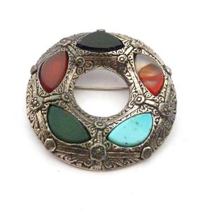 May include: A silver brooch with a circular design, featuring colourful gemstones. The brooch has a central opening and is adorned with red, green, black, and turquoise stones. The metalwork has an intricate, floral pattern.