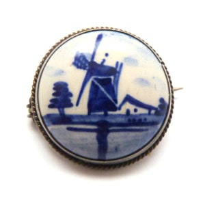 May include: A blue and white porcelain brooch depicting a windmill with a house in the background. The brooch is set in a silver tone metal frame.