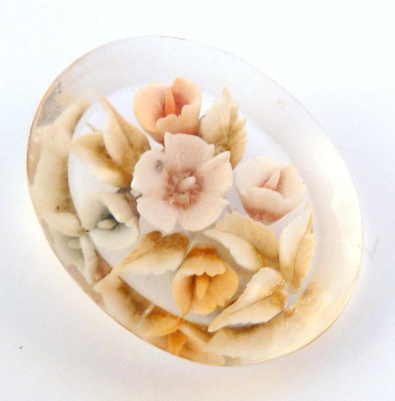 May include: Oval shaped clear resin brooch with a floral design. The flowers are in shades of pink and peach with brown leaves.
