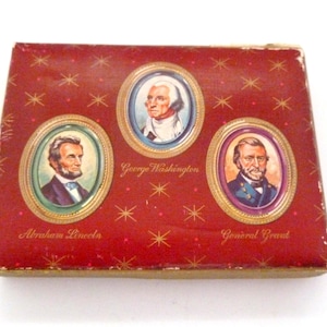 Könnte beinhalten: Eine rechteckige Vintage-Box mit rotem Hintergrund und goldenem Sternmuster. Sie zeigt ovale Porträts von Abraham Lincoln, George Washington und General Grant. Die Porträts sind goldgerahmt, mit den Namen jeder Figur darunter.