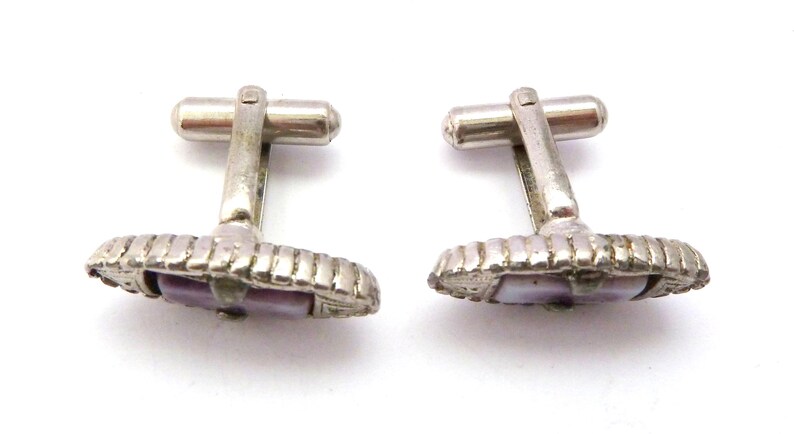 Pu&ograve; includere: Un paio di gemelli d'argento con un'intarsio di madreperla di abalone viola. I gemelli hanno un design decorativo attorno alla conchiglia.