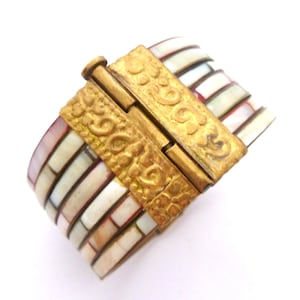 May include: A wide bracelet with rectangular mother-of-pearl segments in white, pink, and cream shades. The bracelet is fastened with a gold-coloured clasp, adorned with intricate embossed patterns. The design of the bracelet resembles a mosaic.