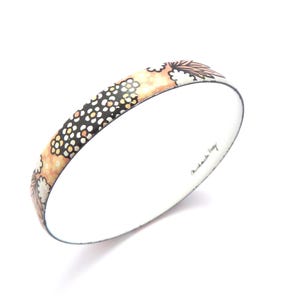 May include: A round bangle bracelet featuring a patterned exterior with black, white, and peach-coloured floral and dotted motifs. The interior of the bracelet is white and displays the signature text Michaela Frey in black script lettering.