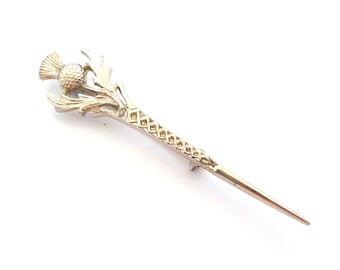 Puede incluir: Un broche de metal dorado con forma de flor de cardo, con una cabeza redondeada y texturizada y hojas puntiagudas. El tallo del broche presenta un intrincado patrón de celosía repetitivo que se estrecha hasta una punta fina y afilada.