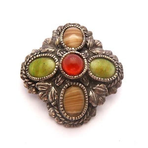 Op de afbeelding: Een vintage broche met een zilverkleurige metalen basis in kruisvorm. Het heeft vier ovaalvormige stenen in bruin- en groentinten, rondom een centrale rode steen. De broche heeft een gedetailleerd, getextureerd ontwerp.