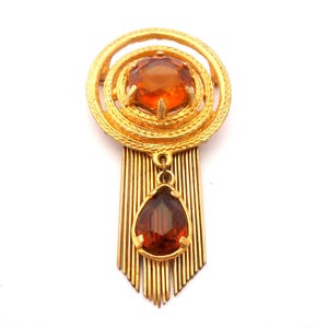 May include: A gold-tone brooch featuring a circular frame with a textured surface and a central round amber-coloured faceted stone. A dangling teardrop-shaped amber-coloured stone hangs from the centre, framed by a series of vertical gold-tone metal rods.