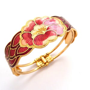 May include: Gold-plated hinged bracelet with a floral design. The bracelet features a large flower with pink and red petals, cream-colored center, and gold accents. The sides have a repeating pattern of red enamel.