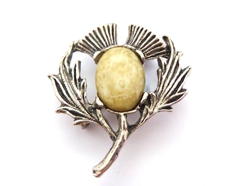 Puede incluir: Broche plateado con forma de cardo, con una piedra ovalada de color amarillo claro en el centro. El broche tiene hojas detalladas y un diseño texturizado, sobre un fondo blanco. Una joya clásica, ideal para chaquetas o bufandas.