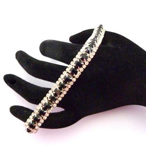 May include: A silver-toned bracelet with black and clear gemstones. The bracelet is set with rows of black stones alternating with rows of clear, sparkling stones. The bracelet is displayed on a black surface.