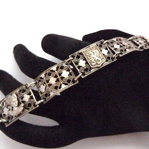 Puede incluir: Pulsera plateada con eslabones de filigrana ornamentados. La pulsera presenta eslabones rectangulares y florales con un diseño central de escudo. La pulsera se muestra sobre una superficie negra.