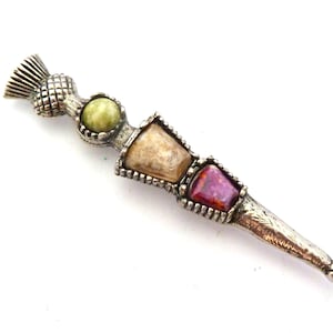 May include: A silver thistle brooch with three inset stones in light green, beige, and purple. The brooch has a textured design and is set against a white background. The brooch is a decorative item.