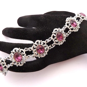 May include: A silver bracelet with oval, magenta gemstones set in decorative floral frames. The bracelet is displayed on a black surface, highlighting its intricate design and vintage style. The bracelet is a piece of jewellery.