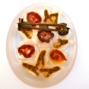 May include: A white, round, glass brooch with a floral design. The flowers are painted in shades of red, brown, and green. The brooch has a metal pin on the back.