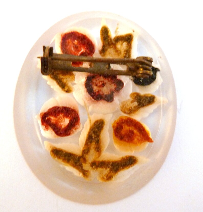May include: A white, round, glass brooch with a floral design. The flowers are painted in shades of red, brown, and green. The brooch has a metal pin on the back.