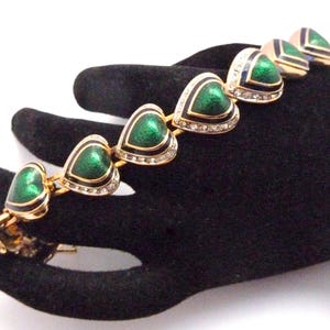 May include: A gold bracelet featuring a series of green enamel heart-shaped charms. Each charm is outlined in black and has a row of clear rhinestones around the edge.
