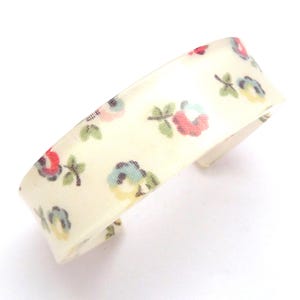 Könnte beinhalten: Ein weißes Stoffarmband mit einem Blumenmuster. Die Blumen sind rot, rosa, blau und grün. Das Armband ist aus einem weichen, flexiblen Material.