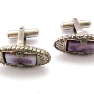 Pu&ograve; includere: Un paio di gemelli d'argento con una pietra viola e bianca incastonata in un design geometrico.