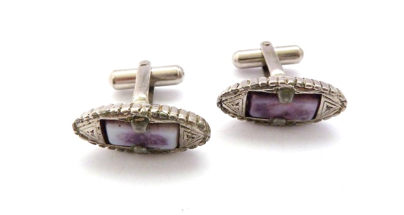 Pu&ograve; includere: Un paio di gemelli d'argento con una pietra viola e bianca incastonata in un design geometrico.