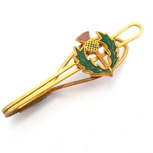May include: Gold-toned tie clip featuring a thistle design with green leaves, a purple flower, and a yellow center. The clip has a classic design, perfect for adding a touch of elegance to any outfit.