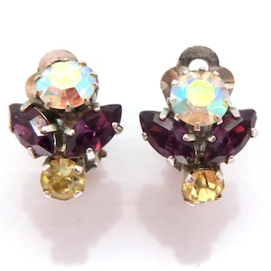 Op de afbeelding: Een paar vintage clip-oorbellen. Elke oorbel heeft een centrale iriserende steen, omringd door dieppaarse en geelgouden stenen, gezet in een zilverkleurig metaal. De oorbellen hebben een bloemmotief.