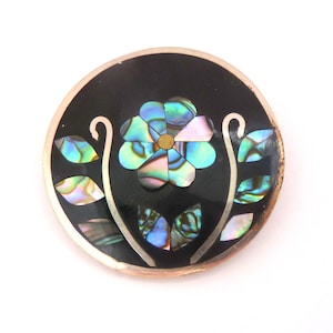 May include: A round, black enamel pin with a silver border. The pin features a blue and green abalone shell flower with a gold center and silver leaves.
