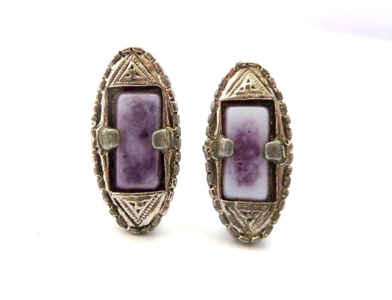 Pu&ograve; includere: Un paio di orecchini d'argento con una pietra rettangolare di colore viola e bianco incastonata in un design geometrico.