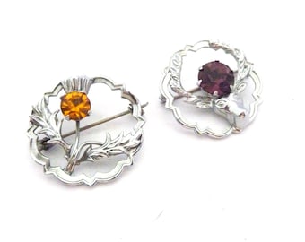 May include: Two silver-toned brooches. One features a thistle with an orange gemstone center, surrounded by leaves. The other has a deer head with a purple gemstone, framed by a wreath. Both have pin backs.