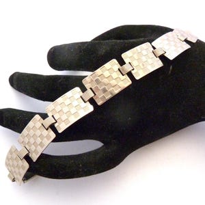 May include: A silver-toned metal link bracelet featuring a series of rectangular plates. Each plate displays a textured checkerboard pattern with alternating matte and polished squares. The rectangular segments are connected by small, ribbed metal bars.