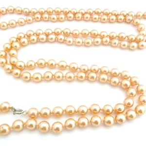 May include: A long necklace featuring numerous peach-toned, spherical beads. The beads are strung together, forming a continuous strand that drapes elegantly. A silver-coloured clasp secures the necklace. The necklace is set against a white backdrop.
