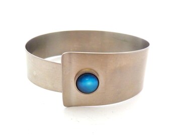 May include: A wide, brushed silver-tone metal cuff bracelet featuring a smooth, rounded band that overlaps at the front. A single, circular, vibrant blue cabochon stone is set securely in the center of the overlapping metal panel.