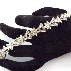 May include: A silver-toned bracelet with a repeating starburst design. The bracelet is set with small, sparkling stones, and is displayed on a black surface. The bracelet is a piece of jewelry.