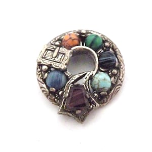 May include: A vintage silver-tone brooch with a circular design. The brooch features a central keyhole shape and is adorned with colourful, round gemstones in shades of orange, green, blue, and turquoise. A small chest is visible on the top.