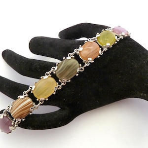 May include: A vintage bracelet featuring oval-shaped stones in shades of brown, yellow, green, and purple, set in a silver-tone metal. The bracelet is displayed on a black hand-shaped display, showcasing its colorful gemstones and intricate design.