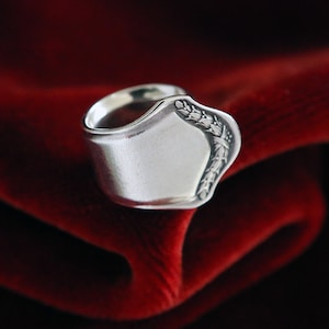Op de afbeelding: Een zilveren ring gemaakt van een hergebruikte lepel met een bloemmotief aan de zijkant. De ring wordt gepresenteerd op een rood fluwelen oppervlak.