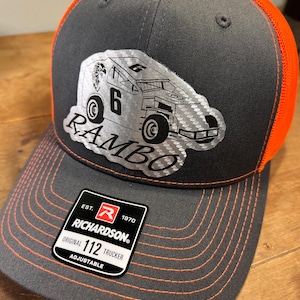 May include: A gray and orange Richardson 112 trucker hat with a silver graphic of a race car and the name "RAMBO". The hat has a curved brim with orange stitching and an adjustable closure.