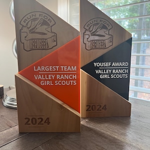 May include: Two wooden award plaques with orange and black accents. The left plaque says "Largest Team Valley Ranch Girl Scouts" and the right plaque says "Yousef Award Valley Ranch Girl Scouts". Both plaques have the text "Run for Refugees 24" and the year "2024" on them.