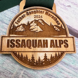 May include: A wooden medal with a brown ribbon. The medal is engraved with the text "Father-Daughter Marathon 2024" and "ISSAQUAH ALPS". The medal features a mountain range and trees.