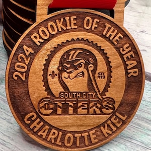 May include: A wooden medal with the text "2024 Rookie of the Year" and "South City Otters Charlotte Kiel" engraved on it. The medal features a cartoon otter wearing a helmet and riding a bicycle.