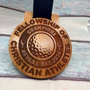 Puede incluir: Medalla de madera con cinta azul marino. La medalla redonda presenta las inscripciones grabadas "FELLOWSHIP OF CHRISTIAN ATHLETES" y "CHAMPIONS NEVADA DAY 2024" alrededor de un diseño de pelota de golf. La medalla es de madera marrón claro.