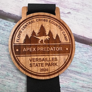 Op de afbeelding: Een houten medaille met een zwart lint. De medaille heeft een dinosaurus silhouet, bomen en de tekst "Floyd Central Cross Country Camp", "Apex Predator", "Versailles State Park" en "2024".