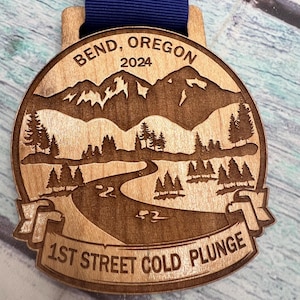 Op de afbeelding: Een houten medaille met een blauw lint. De medaille toont een berglandschap met een rivier die er doorheen slingert. De tekst "Bend, Oregon 2024" staat bovenaan de medaille. De tekst "1st Street Cold Plunge" staat onderaan de medaille.