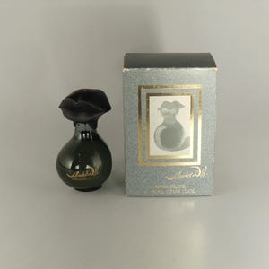 May include: A dark green perfume bottle with a black lip-shaped cap and gold lettering, next to a matching box. The box features a framed image of the bottle and the words "Salvador Dali" and "After Shave".