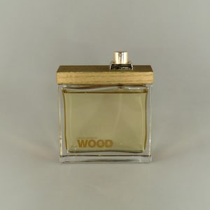May include: A rectangular glass perfume bottle with a wooden cap and a gold-coloured sprayer. The bottle contains a light amber liquid and the word "WOOD" is printed on the front. The bottle is on a light grey background.