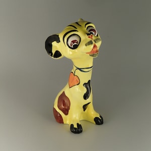 Peut inclure: Une figurine en céramique jaune d'un chien avec des marques noires et brunes. Le chien a de grands yeux, une moustache noire et un cœur rouge sur la poitrine.