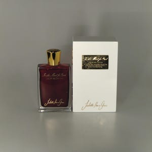 Op de afbeelding: Een glazen fles parfum met een gouden dop en een rode vloeistof. De fles is gelabeld met "La Plus Belle" en "Juliette Has a Gun". De fles staat op een wit oppervlak naast een witte doos met hetzelfde label.